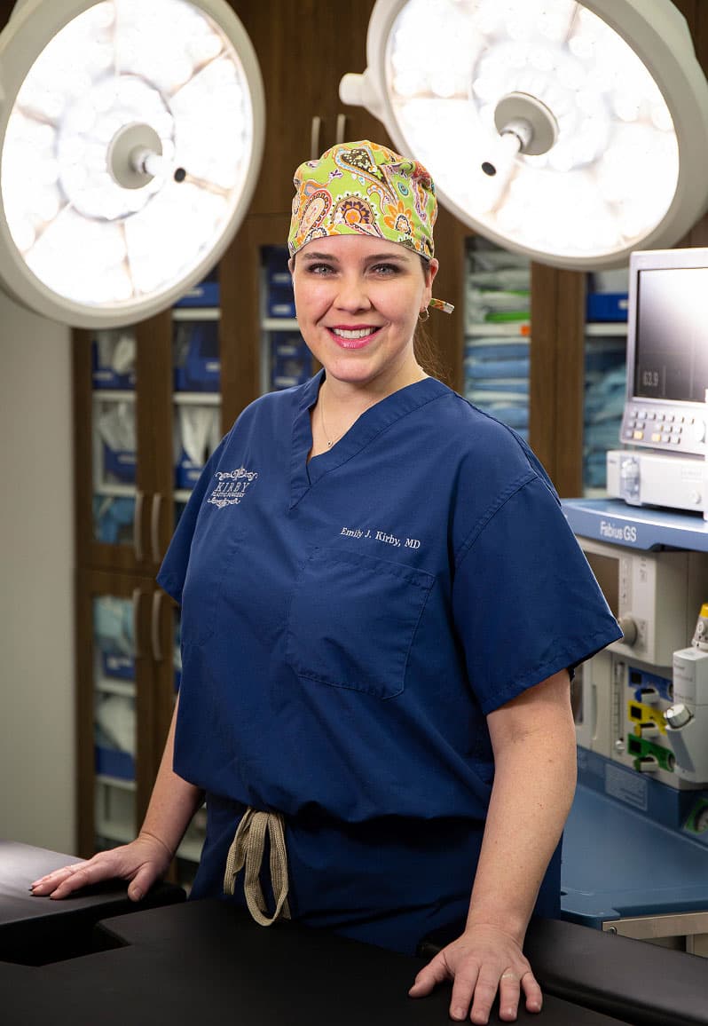 Female plastic surgeon Dr. Emily J. Kirby stands under bright LED lights wearing scrubs in the accredited, state-of-the-art, on-site operating room at the Fort Worth plastic surgeon's practice