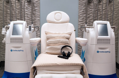 A white reclining treatment chair with pillows, blanket, and a headset, located next to two CoolSculpting machines, which are able to sculpt two fat deposits at once (also known as DualSculpting). CoolSculpting is a non-surgical fat reduction treatment that uses cool temperatures to dissolve unwanted fat deposits in areas like the abdomen and flanks (love handles).